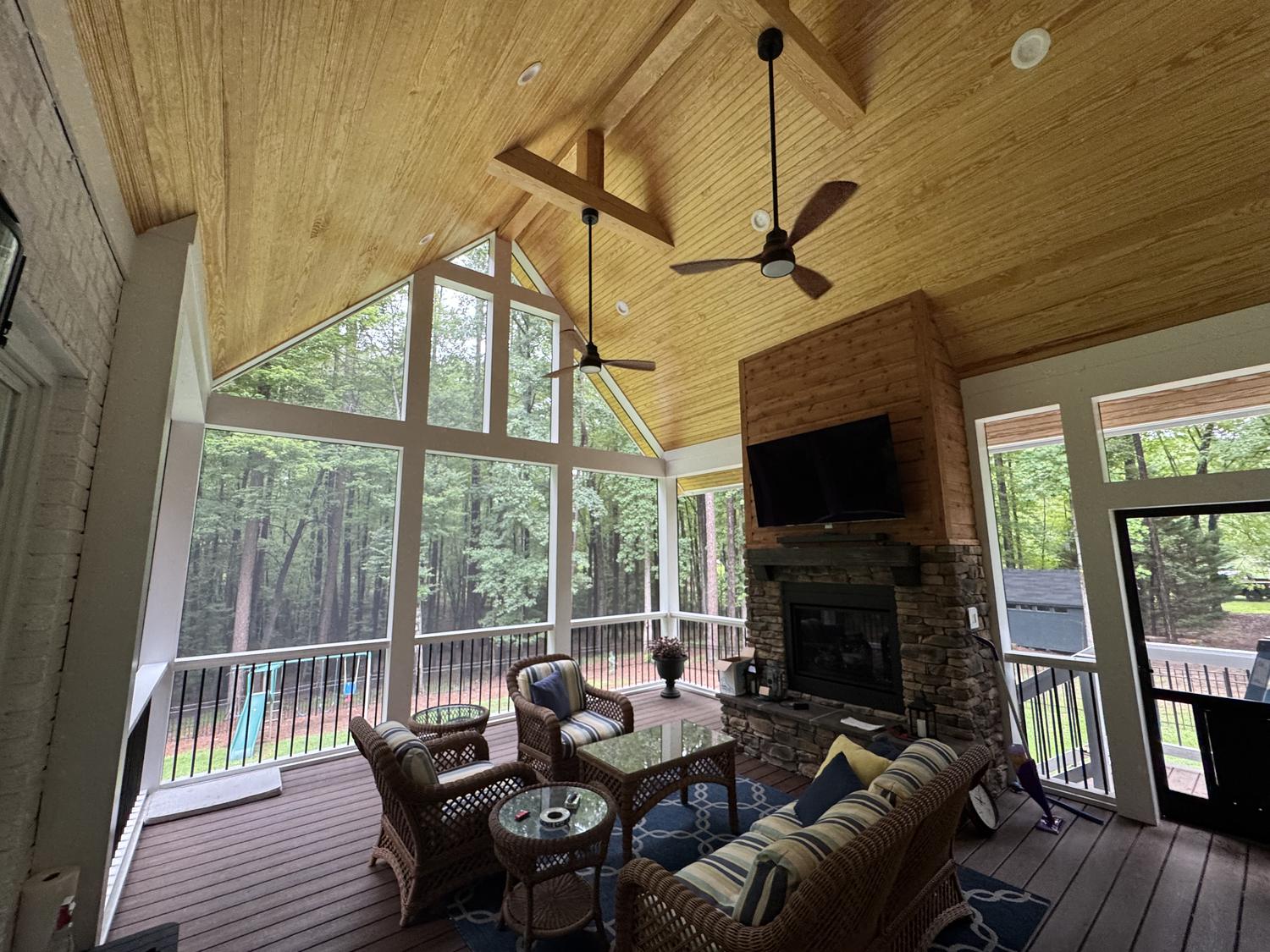 A completed screened porch with a vaulted tongue-and-groove wood ceiling, exposed beams, a stone fireplace, and floor-to-ceiling screens overlooking the woods