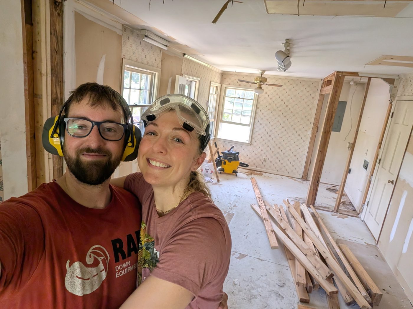 A couple in safety glasses and hearing protection mid-demolition, standing in a gutted room with framing exposed